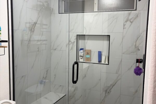 Walk-in shower remodel with marble-look tile, built-in bench, niche, frameless glass door and black hex floor by Western Construction & Restoration